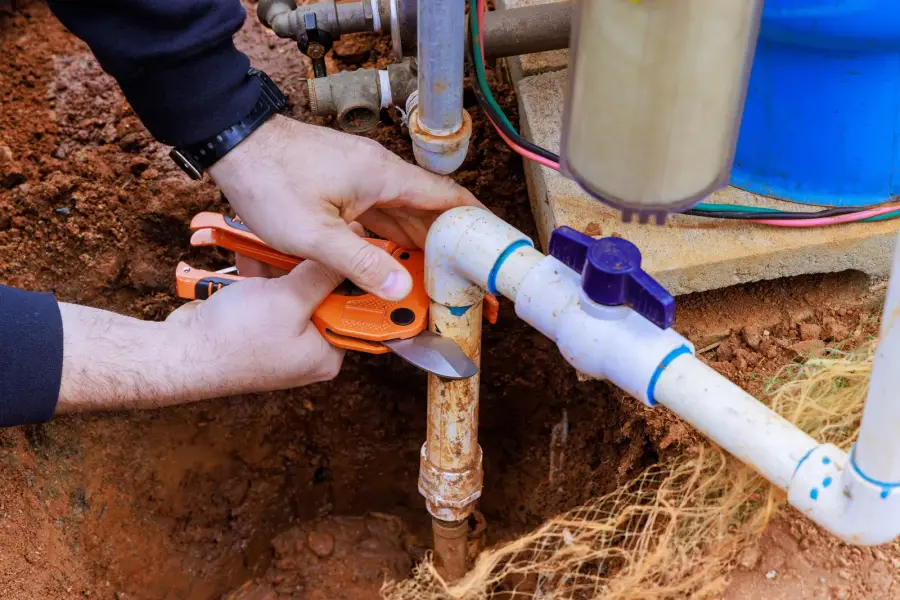 Plumber's gloved hands cutting into a corroded water supply pipe beside a shut-off valve in an excavated trench